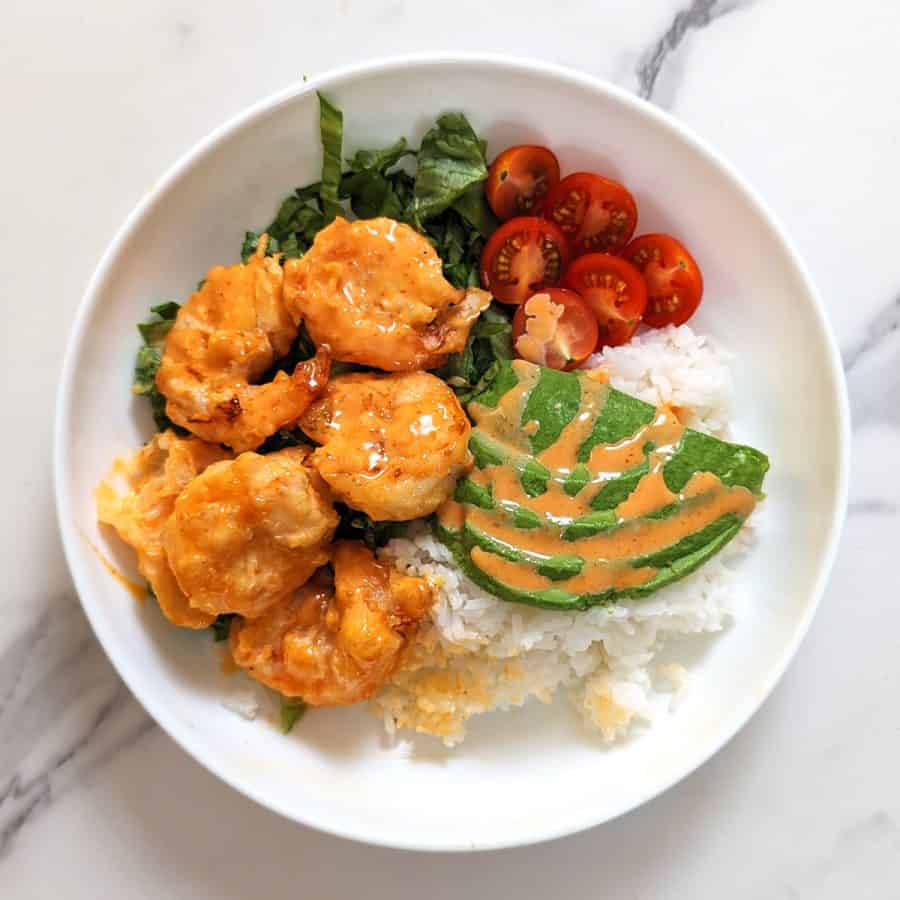 A white plate with air-fried shrimp on top of a bed of shredded lettuce and white rice with a side of sliced cherry tomatoes and avocados, topped with honey sriracha drizzle