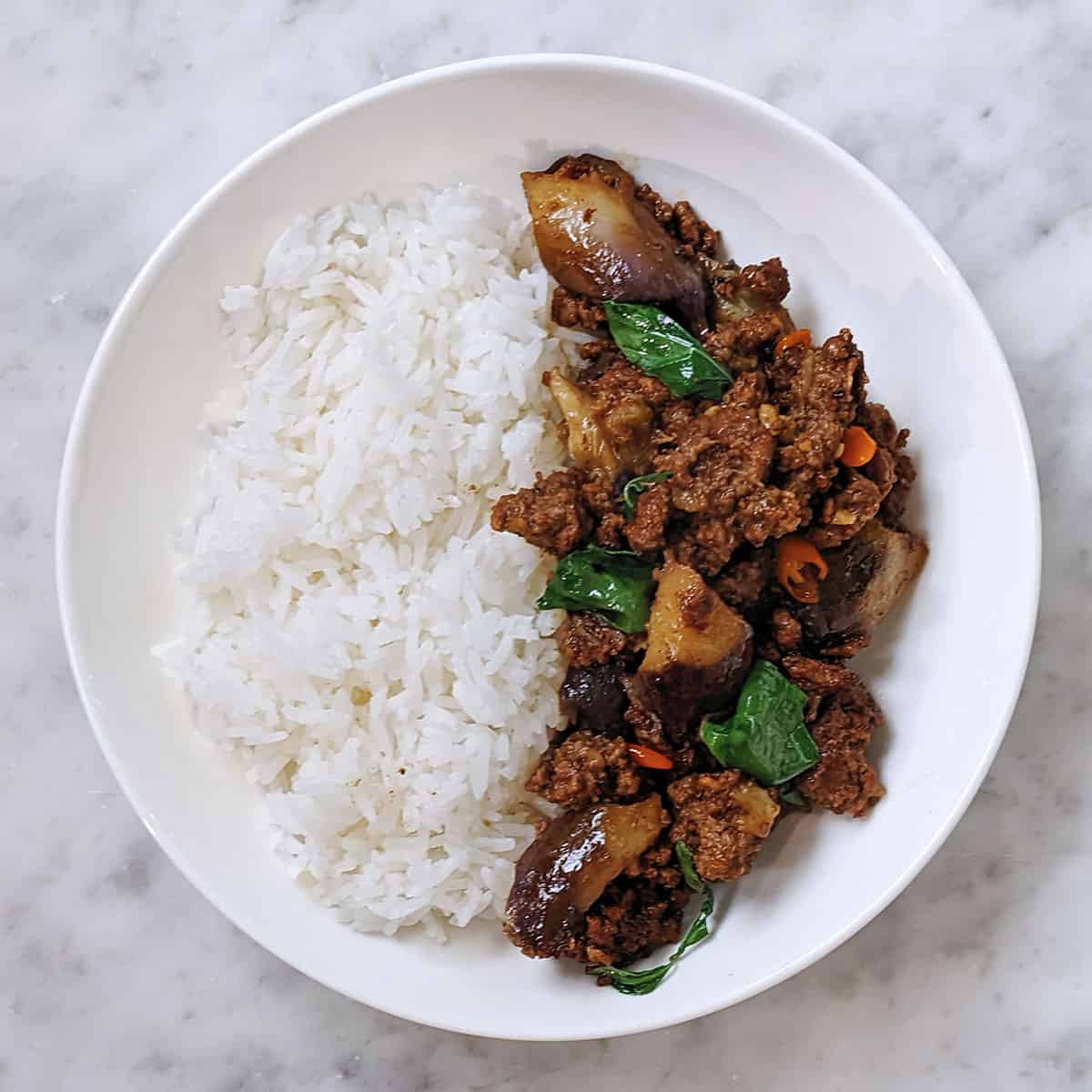 A plate half full with steamed white rice on one side and thai basil beef and eggplant on the other on a marble counter top