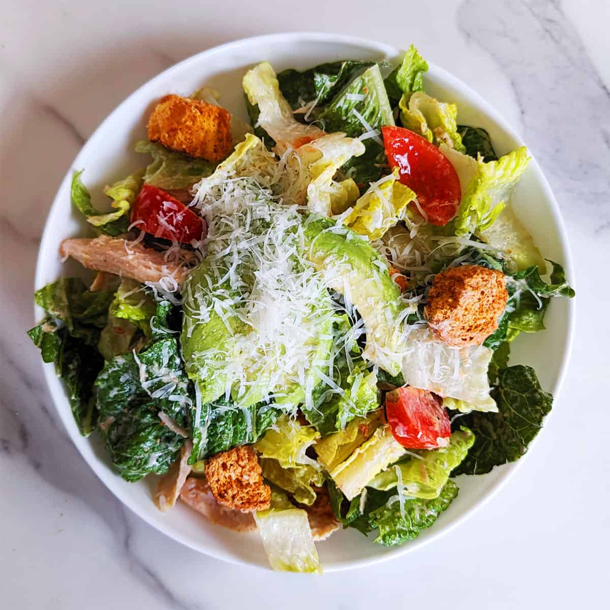A plate of Caesar salad topped with avocado and shredded parmesan on a marble countertop