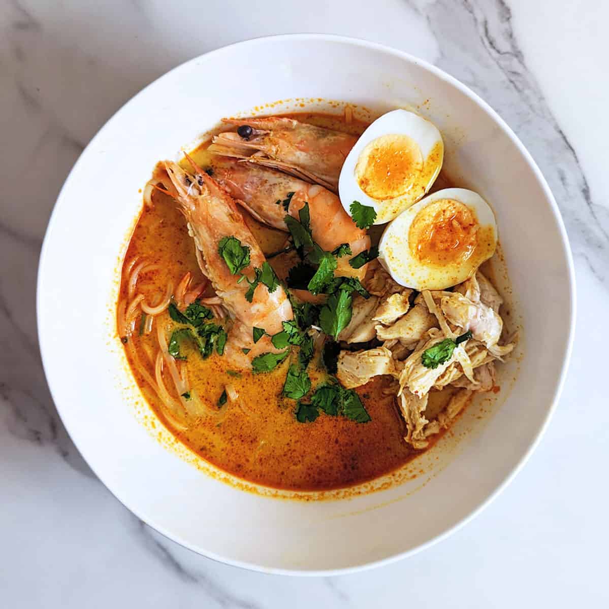 A bowl of chicken curry laksa, topped with hard-boiled eggs, shrimp, and cilantro on a marble countertop