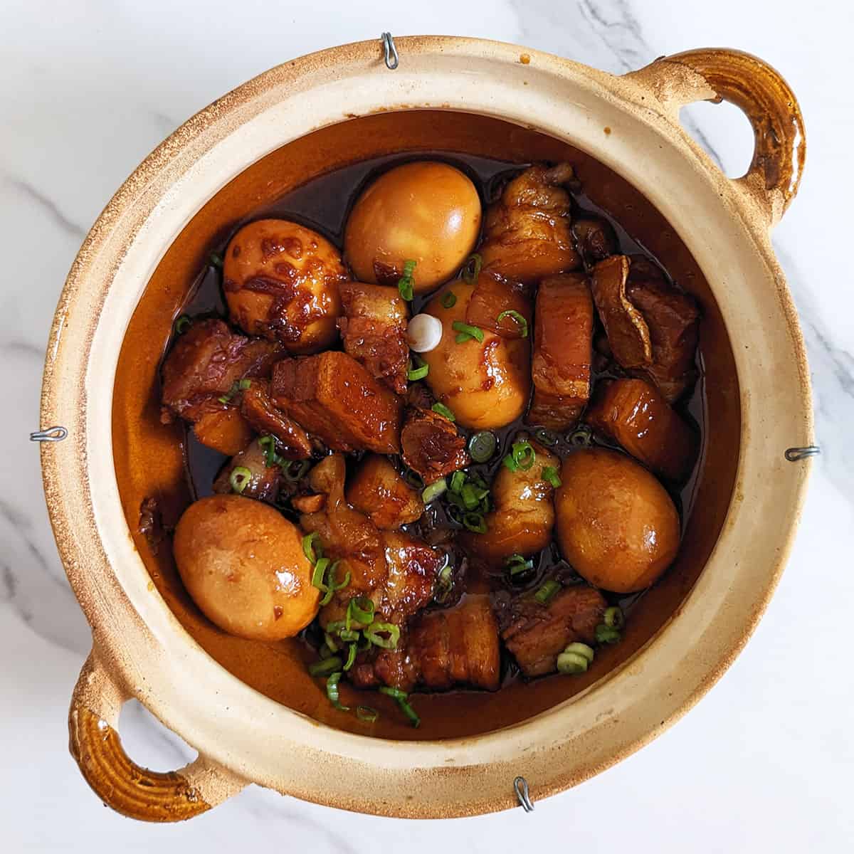 A photo of a claypot containing soy sauce braised pork belly with hardboiled eggs on top of a marble backdrop