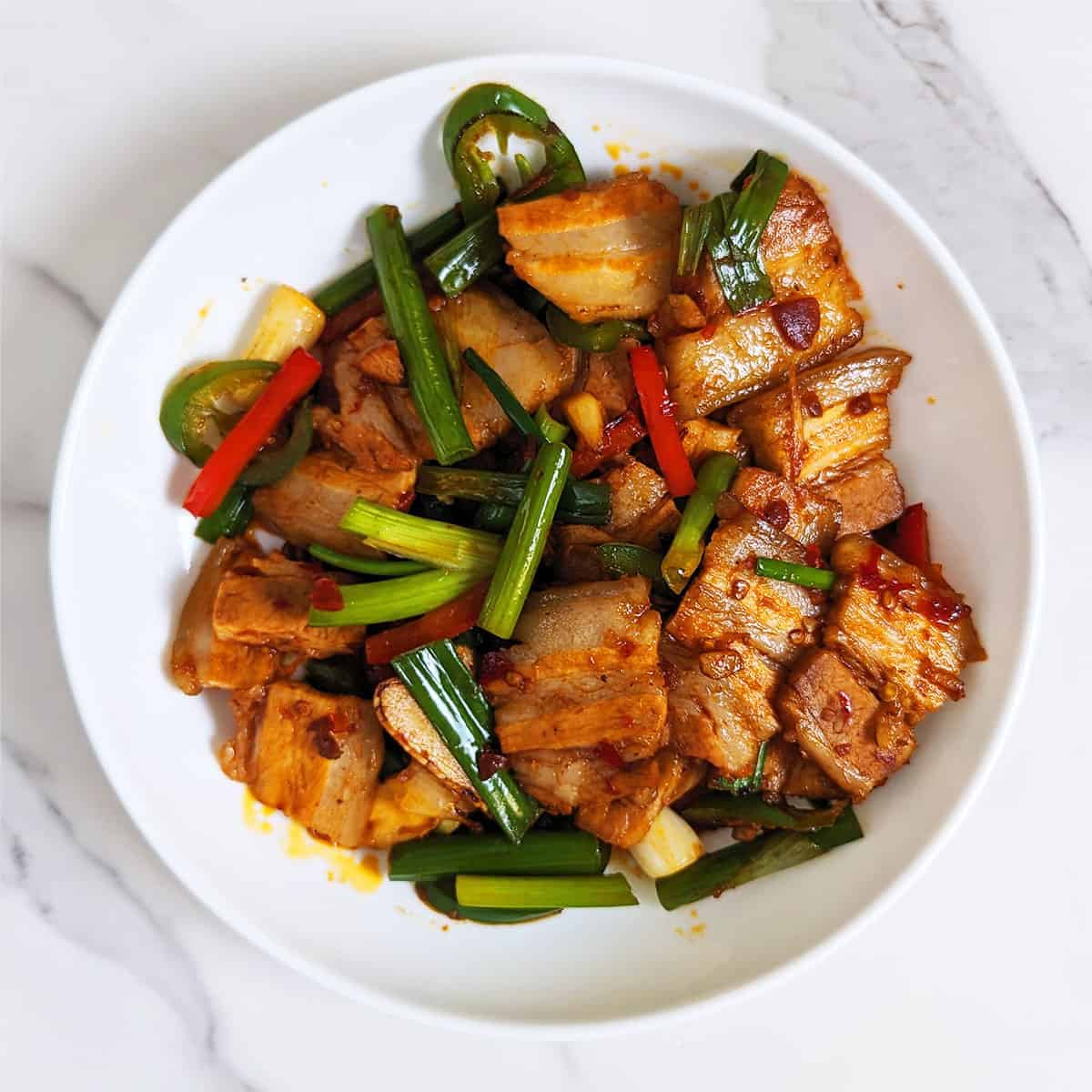 A plate of stir-fried pork belly with julienned red bell pepper and chopped green onions on a marble backdrop