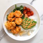 A white plate with air-fried shrimp on top of a bed of shredded lettuce and white rice with a side of sliced cherry tomatoes and avocados, topped with honey sriracha drizzle