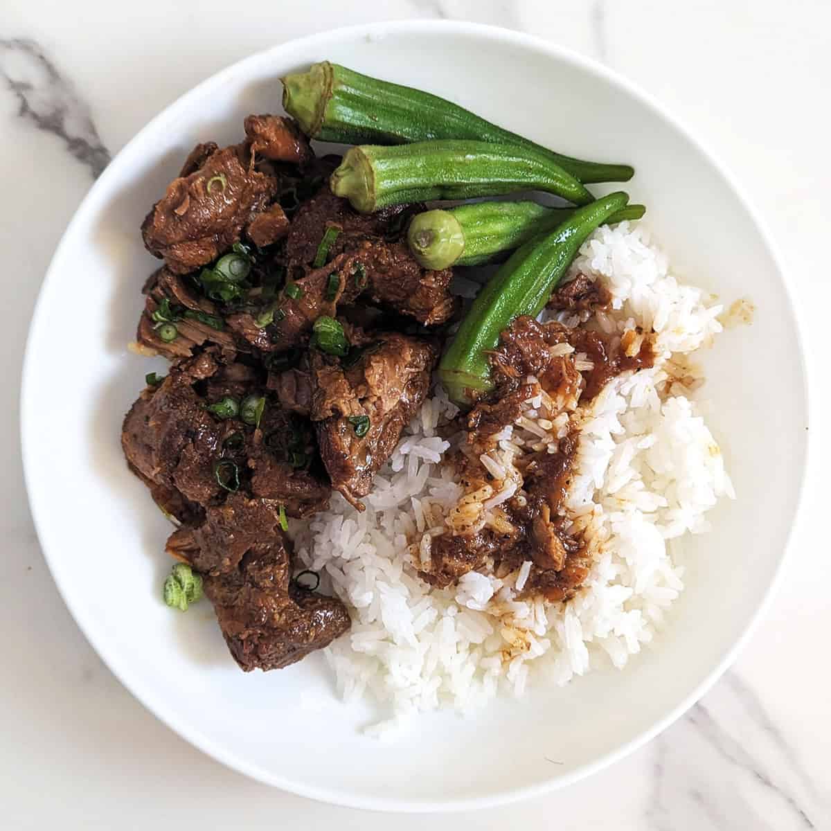 A plate of Filipino pork adobo with steamed rice and okra on a while marble background